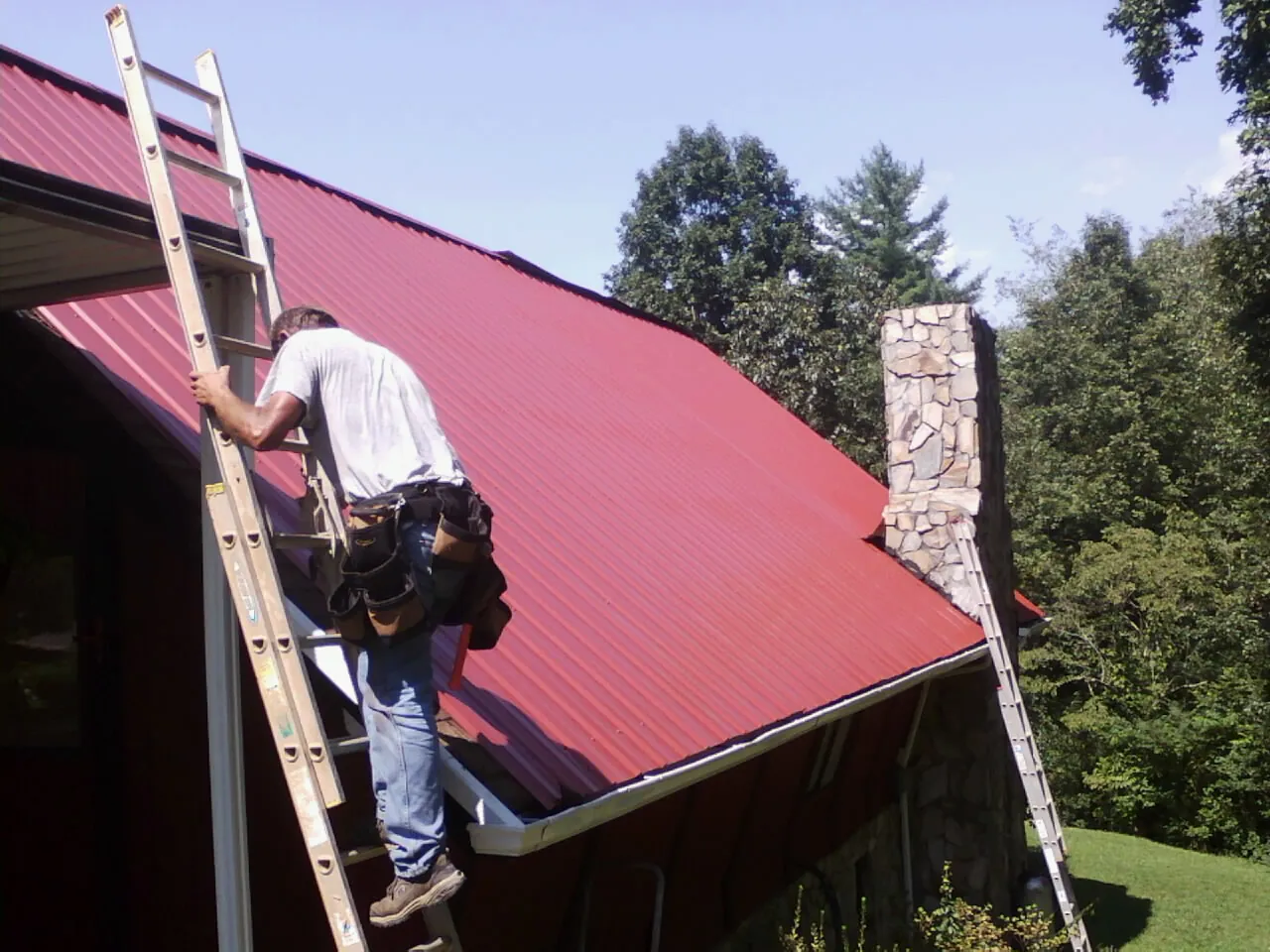 Roofer on a ladder beside a red metal roof and stone chimney — Soffit & Fascia Repair by Roofing Brothers in Olney Springs